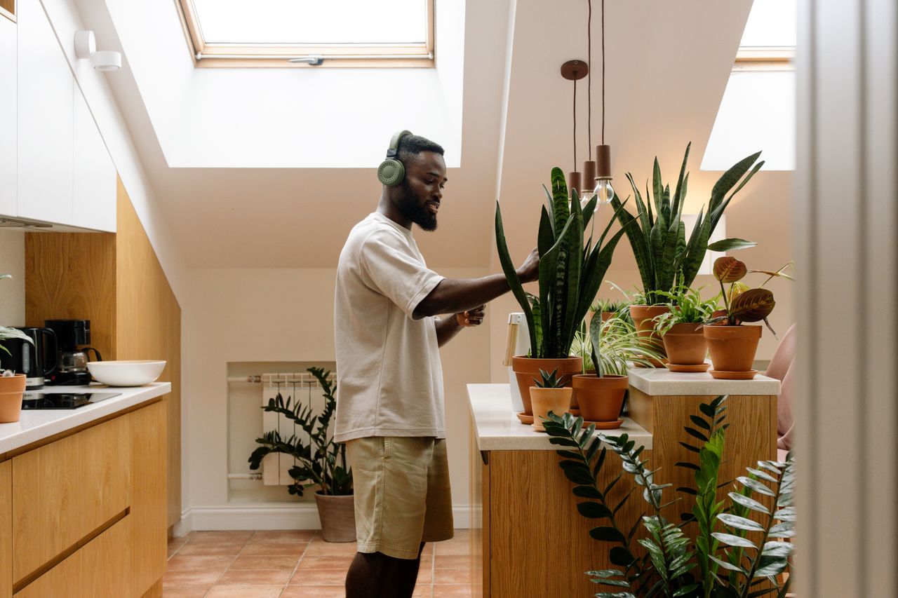 A man listening to music with headphones while watering indoor plants in a modern kitchen.